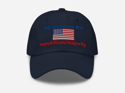 A navy dad hat with the embroidered text "my pronouns are republican/majority" in bold red and blue, featuring a detailed American flag design at the center, viewed from the front